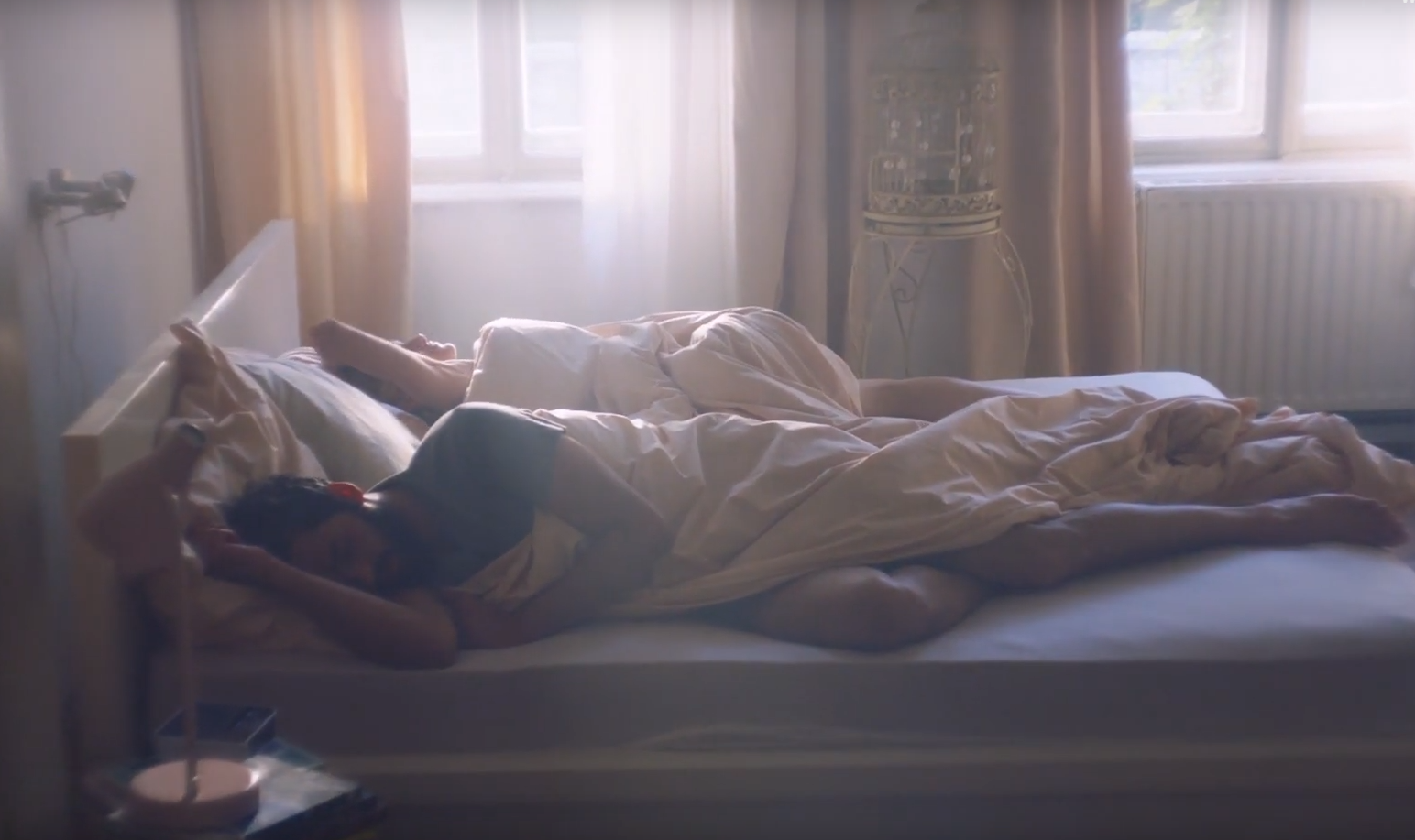 Na sliki sta moški in ženska, ki spita v zakonski postelji, zavita v odejo, ob jutranji svetlobi skozi okno.