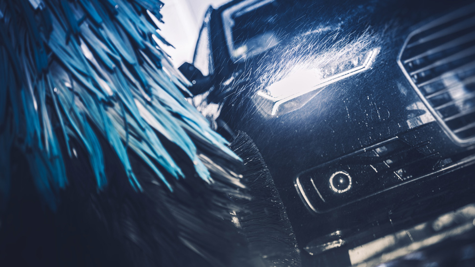 Na sliki je sprednji del avtomobila z vključenimi žarometi v avtopralnici. Modre krtače in curek vode čistijo površino vozila.