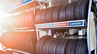 Na sliki so avtomobilske gume zložene na kovinskih policah v trgovini. Nad gumami je napis Petrol eSHOP