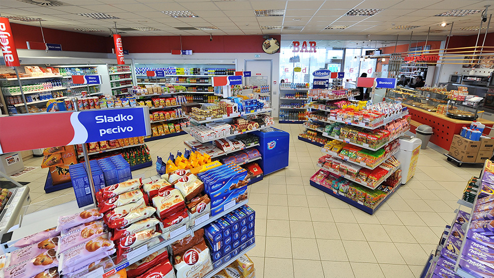 Notranjost Petrolove trgovine HipShop s policami, polnimi prigrizkov, pijač in osnovnih živil. Na oznakah so razdelki, kot je 'Sladko pecivo'.