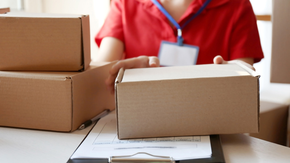 Na sliki je oseba v rdeči uniformi s pripeto identifikacijsko kartico, ki ravna s kartonskimi paketi, položenimi na mizo