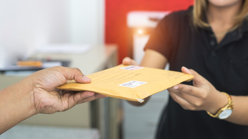 Na sliki sta dve osebi, ena predaja rumeno kuverto drugi, ki jo sprejema. V ozadju je pisarniško okolje.