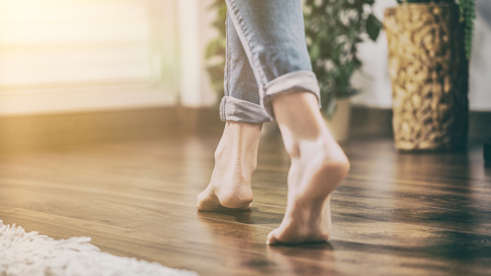 Na sliki so bose noge osebe, ki hodi po lesenem parketu v domačem ambientu. V ozadju sta vidni okrasni lončnici.