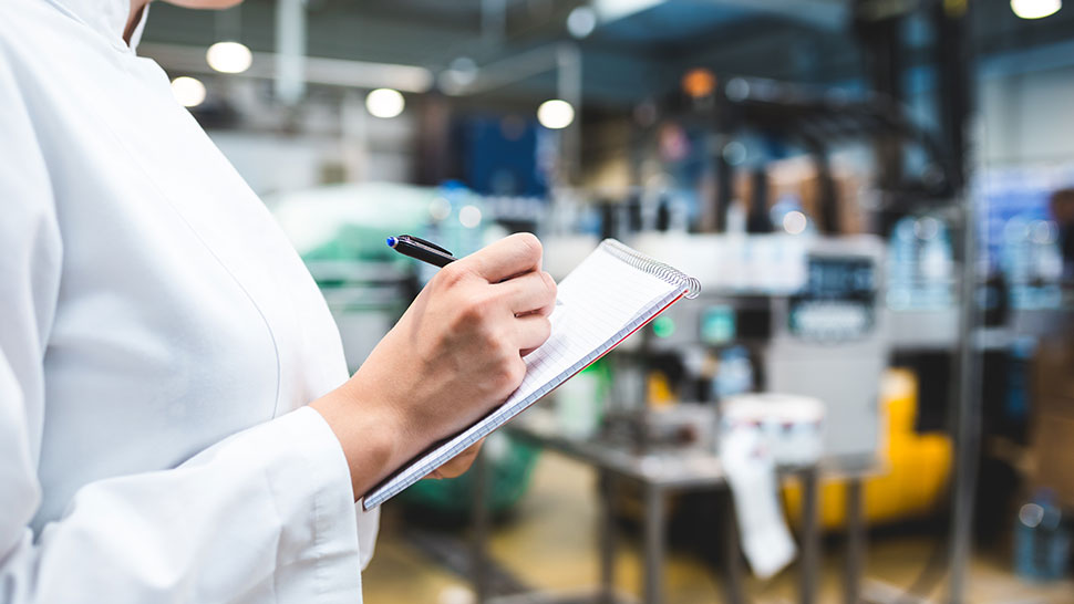 Na sliki je oseba v belem laboratorijskem plašču, ki drži pisalo in zapisuje v beležko. V ozadju je zamegljen industrijski oziroma proizvodni prostor, kjer se vidijo različni stroji, oprema in materiali.