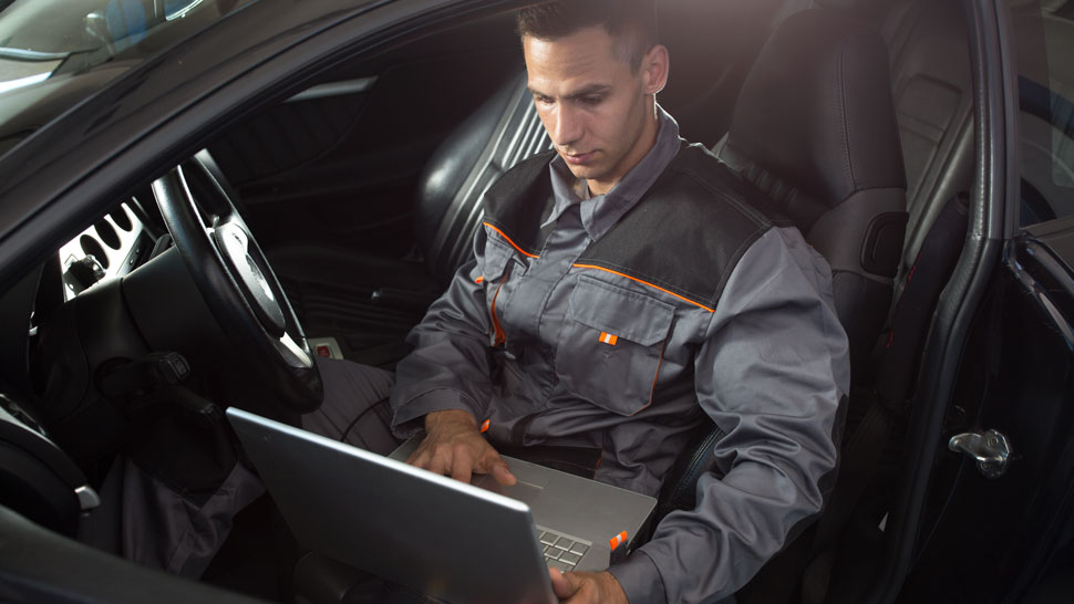 Na sliki je mehanik, ki sedi v vozilu in dela na prenosnem računalniku. Oblečen je v delovno obleko in očitno izvaja diagnostiko ali pregled vozila s pomočjo računalniške programske opreme.