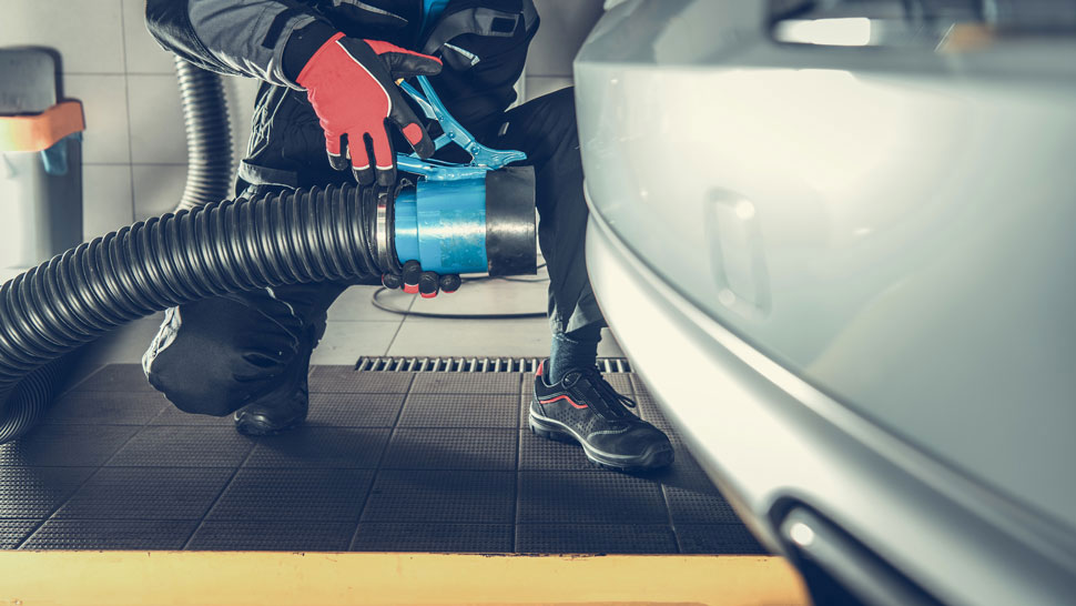 Na sliki oseba v delovni obleki in rokavicah priključuje velik sesalni cevni sistem na izpuh avtomobila. Postopek poteka v delavnici ali servisnem prostoru za namen merjenja emisij vozila.