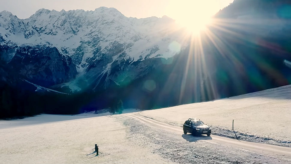 Na sliki je avtomobil, ki vozi po zasneženi cesti v čudoviti gorski pokrajini. V ozadju se dvigajo mogočne zasnežene gore, sonce pa sije močno nad njimi in ustvarja svetlobne žarke.