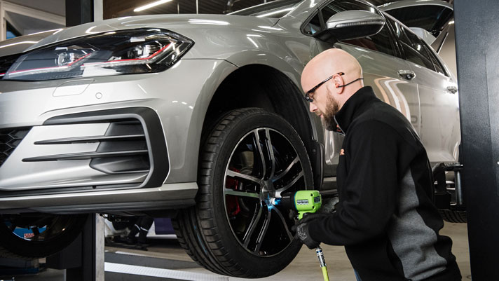 Na sliki je avtomehanik, ki s strojem privijača vijake na kolesu avtomobila. Srebrn avto je dvignjen na dvigalu v delavnici.