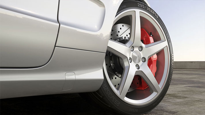 Na sliki je spodnji del belega avtomobila, v ospredju je platišče s petimi kraki in rdečimi zavorami. Vozilo stoji na asfaltirani površini.