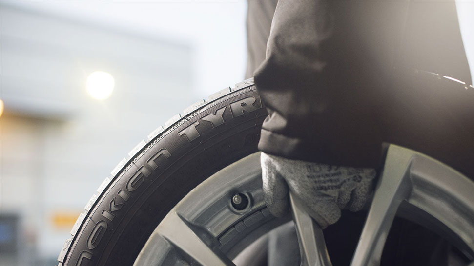Na sliki je oseba z delovno rokavico, ki drži avtomobilsko kolo z napisom Nokian Tyres na pnevmatiki. V ospredju je jasno viden rob platišča in profil gume.