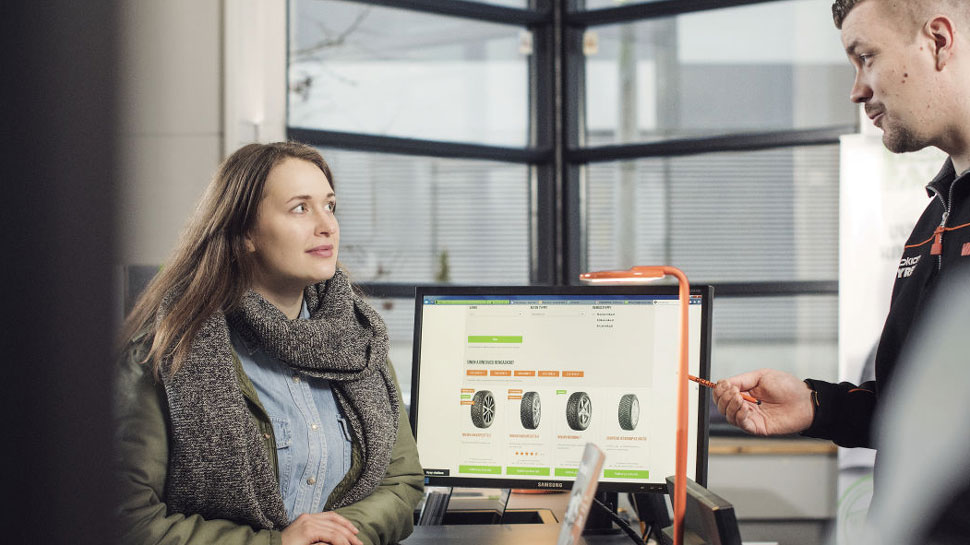 Na sliki je ženska pri pultu, kjer se s prodajalcem pogovarja o nakupu pnevmatik. Na računalniškem zaslonu sta prikazana izbor in cene gum.