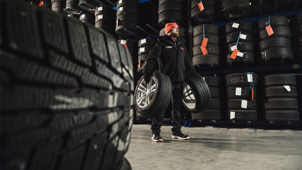 Na sliki je delavec v skladišču gum, ki v rokah nosi dve avtomobilski kolesi. Okoli njega so police, polne zloženih pnevmatik.