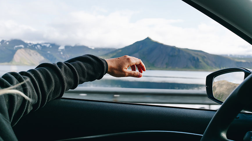 Na sliki je notranjost vozila, iz katere oseba iztegne roko in jo nasloni na vrata ter čez okno avtomobila. V ozadju se odpira čudovita pokrajina s hribi oziroma gorami ob vodi.