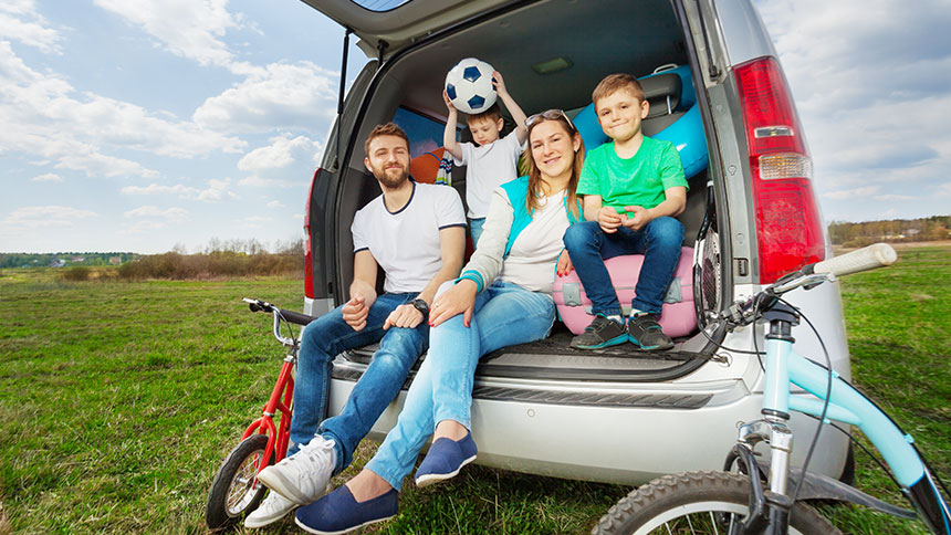 Na sliki je mlada družina na izletu. Sedijo na prtljažniku srebrnega avtomobila, parkiranega na travniku. Starša in dva otroka uživajo na prostem – eden od otrok drži žogo. Ob avtomobilu sta naslonjena dve otroški kolesi.