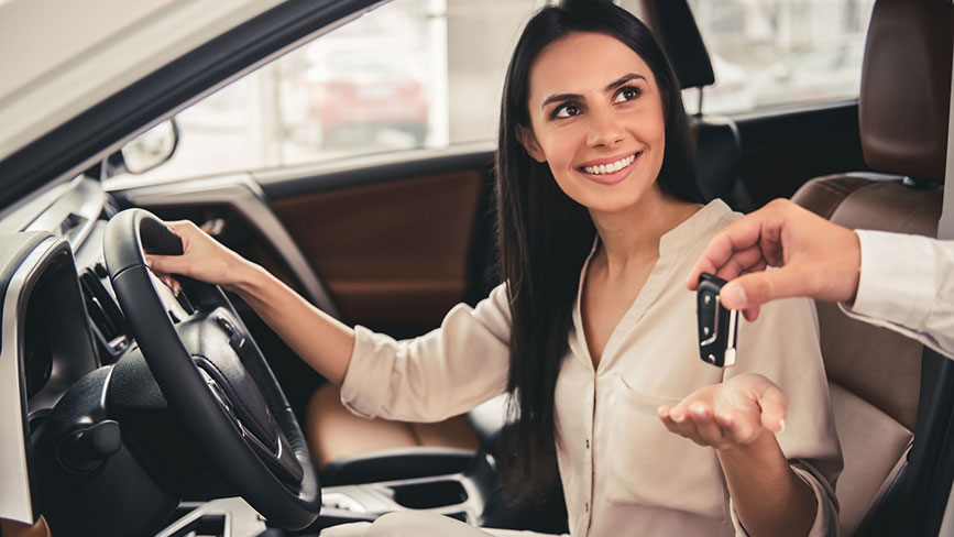 Na sliki je ženska v avtomobilu, ki sedi na voznikovem sedežu in se nasmejano ozira proti osebi zunaj vozila. Nekdo ji izroča ključe od avtomobila, ki jih z veseljem sprejema v roko. V ozadju je notranjost avtomobila v svetlo-rjavih tonih.