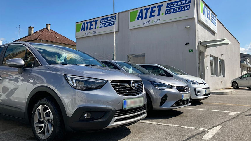 Na sliki je parkiranih več vozil pred poslovalnico podjetja ATET rent a car. Vidni so različni avtomobili (na čelu s sivo Opelovo vozilo), v ozadju pa je stavba z več logotipi podjetja ATET in kontaktnimi podatki.