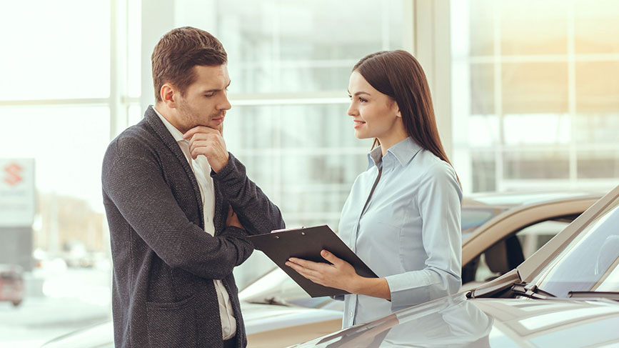 Na sliki sta moški in ženska v avtohiši. Ženska v svetli srajci drži mapo in nekaj razlaga moškemu, ki zamišljeno posluša in se z roko dotika brade. Oba stojita ob avtomobilu, v ozadju pa je svetel razstavni prostor z več vozili.