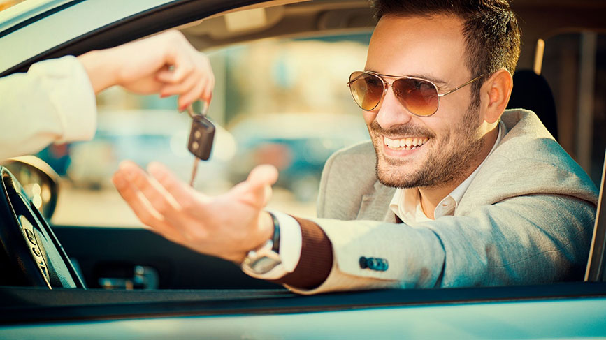 Na sliki je moški v avtomobilu, ki z nasmehom in iztegnjeno roko prejema avtomobilske ključe. Oblečen je elegantno, nosi sončna očala in deluje samozavestno ter zadovoljen. Prizor izraža občutek zaupanja, udobja in zadovoljstva ob prevzemu vozila.