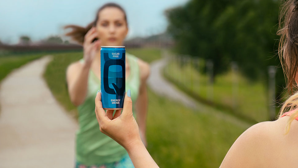 Ženska v športni opravi drži pločevinko modrega Q Energy Drink Sugar Free proti kameri, v ozadju pa po stezi proti njej teče druga ženska. Slika je posneta na prostem, ob tekaški poti z zelenjem.