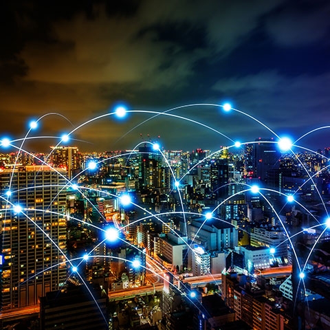 slika prikazuje : Nočno mestno jedro z osvetljenimi stavbami in svetlobnimi linijami, ki ponazarjajo digitalno povezljivost in omrežja