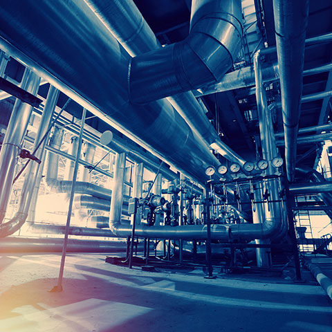 Slika prikazuje notranjost industrijskega objekta z velikimi kovinskimi cevmi, merilniki tlaka in prezračevalnimi kanali.