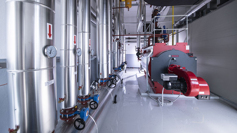 Na sliki je prikazana kotlovnica z velikim industrijskim kotlom in več kovinskimi cevmi z merilniki tlaka.