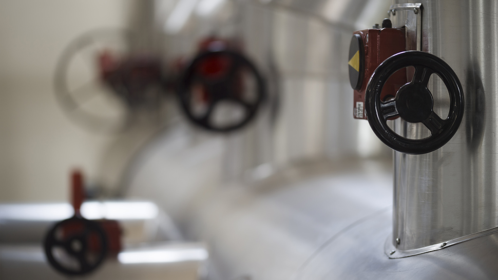 Slika prikazuje industrijski cevovod z več kovinskimi cevmi in rdečimi ventili za uravnavanje pretoka.