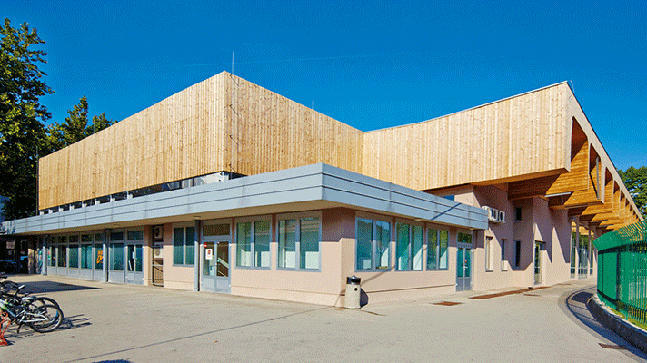 Slika prikazuje manjše slovensko naselje z rdečimi strehami, obdano z zelenimi travniki in gozdnatimi hribi v ozadju.