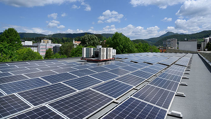 Na sliki je prikazana streha z nameščenimi sončnimi celicami in prezračevalnimi napravami; v ozadju so drevesa, stanovanjske stavbe in hribovita pokrajina pod modrim nebom.