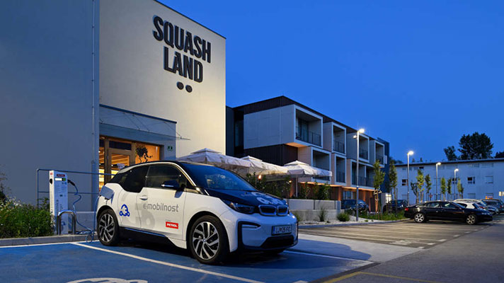 Na sliki je prikazana polnilna postaja pred stavbo Squash Land, kjer je parkiran bel električni avtomobil z nalepljenimi logotipi.