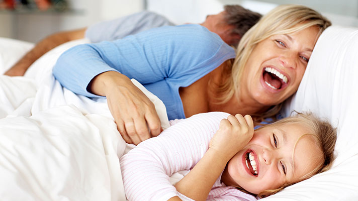 Na sliki je nasmejana družina v postelji – ženska in otrok v ospredju se smejita, v ozadju pa leži moški.