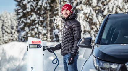 Moški v rdeči kapi Petrol pozira ob električni polnilnici v zasneženem gozdu in drži polnilni kabel, zraven je parkiran električni avto.