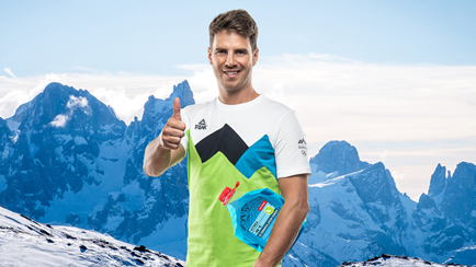 Moški v slovenski reprezentančni majici stoji v zasneženih gorah, v eni roki drži kanister hladilne tekočine, z drugo kaže dvignjen palec.