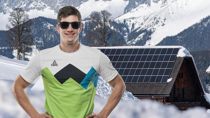 Moški v slovenski reprezentančni majici in sončnih očalih stoji v zasneženi pokrajini pred hišo s sončno elektrarno na strehi.