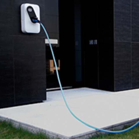 Na sliki je domača električna polnilnica, nameščena na temni fasadi sodobne hiše. Iz polnilnice vodi moder kabel, ki je priključen na električno vozilo zunaj kadra.