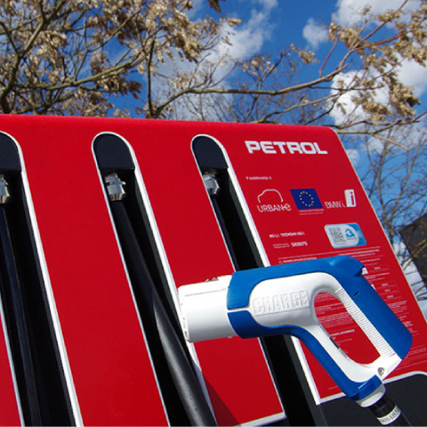 Na sliki je Petrolova električna polnilnica z rdečim ohišjem, na kateri je vidna modro-bela polnilna ročka z napisom »CHARGE«. Na polnilnici so logotipi Evropske unije, programa URBANE in EBRD.