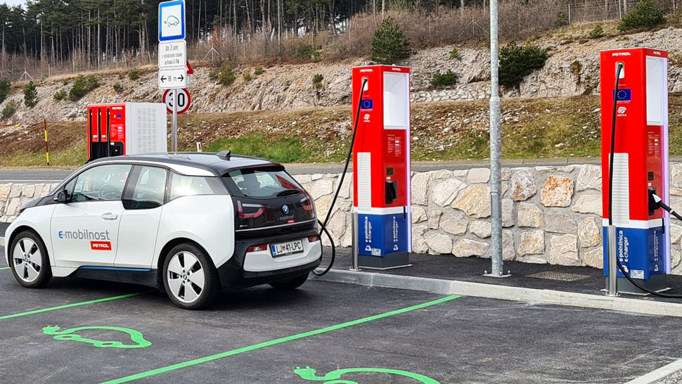 Na sliki je električno vozilo BMW i3 z oznako e-mobilnost Petrol, ki se polni na Petrolovi električni polnilnici. Polnilnici sta postavljeni ob kamnitem zidu, označeni v rdeče-modri barvni kombinaciji, okoli vozila pa so na tleh vidne zelene talne oznake za parkirna mesta namenjena električnim avtomobilom.