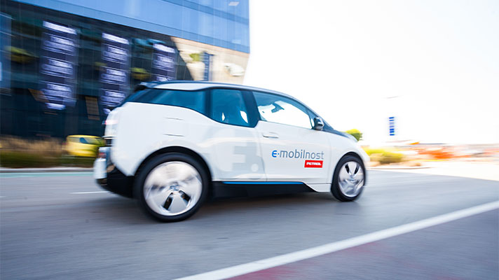 Na sliki je električni avtomobil BMW i3 z oznako e-mobilnost Petrol, ki se pelje po cesti. Avto je bel z modrimi in črnimi poudarki.