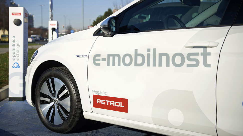 Na sliki je bel električni avtomobil z napisom “e-mobilnost” in logotipom Petrol, parkiran ob električni polnilnici Petrol e-charger. Vozilo je prikazano od strani, v ospredju pa izstopajo napisi in dizajn na vratih avtomobila.