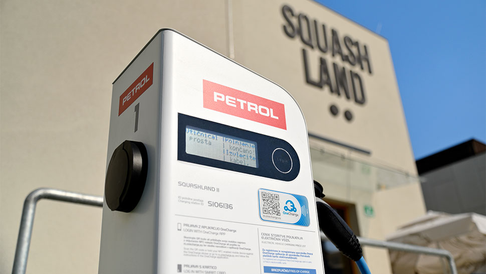 a sliki je Petrolova električna polnilnica postavljena pred objektom z napisom Squash Land. Polnilnica je bele barve z rdečim logotipom Petrol, na zaslonu pa so prikazane informacije o stanju priključkov in polnjenju. Na sprednji strani je tudi nalepka z oznako e-polnilnica in QR kodo.