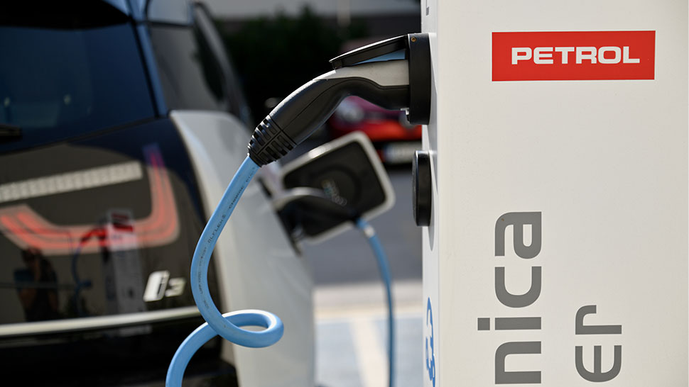 Na sliki je Petrolova električna polnilnica bele barve z rdečim logotipom Petrol. V ospredju je vidno priključeno modro polnilno kabelsko povezavo, v ozadju pa je parkirano električno vozilo, ki se polni.
