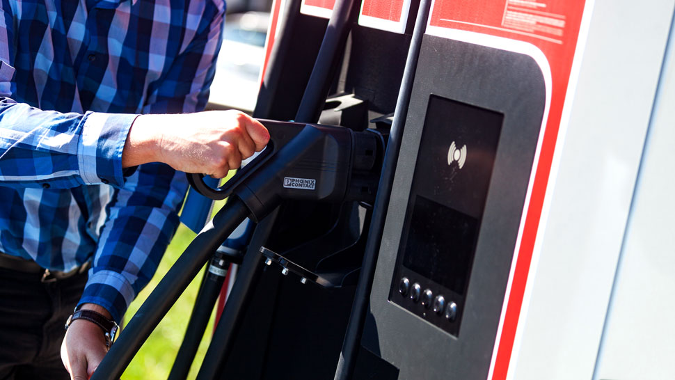 Na sliki oseba v modri karirani srajci priključuje kabel za polnjenje na Petrolovo električno polnilnico v rdeče-beli barvni kombinaciji. V ospredju je razviden zaslon polnilne postaje in priključek, medtem ko oseba z eno roko drži in priklaplja polnilni kabel.