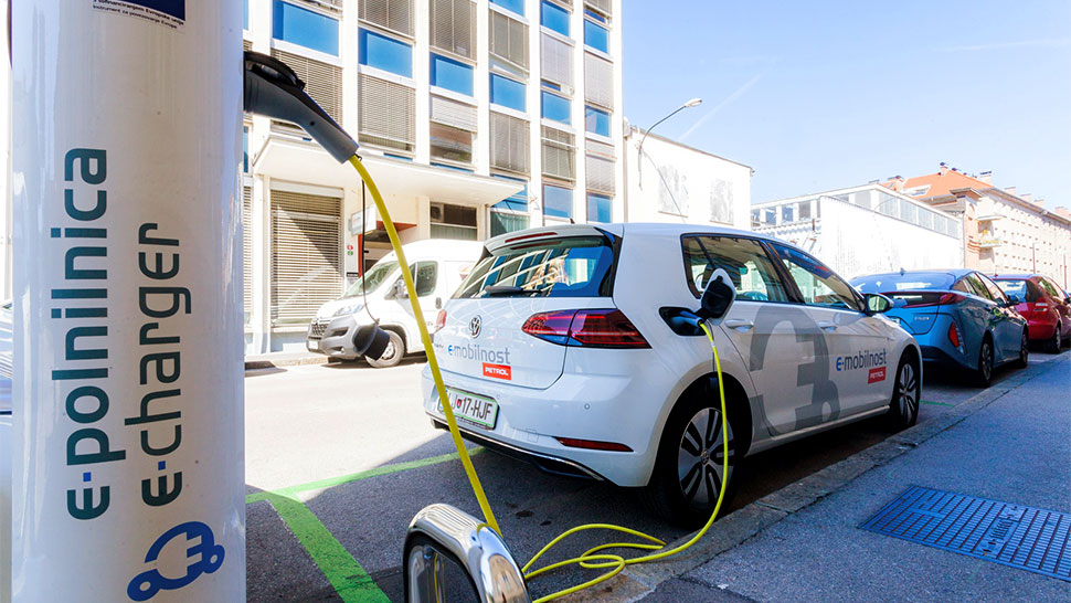 Na sliki je Petrolova e-polnilnica z napisom e-polnilnica / e-charger, ob kateri je parkiran električni avtomobil z oznako e-mobilnost Petrol. Avto je povezan s rumenim kablom za polnjenje, okolica pa prikazuje mestno ulico z več parkiranimi vozili in poslovnimi stavbami.