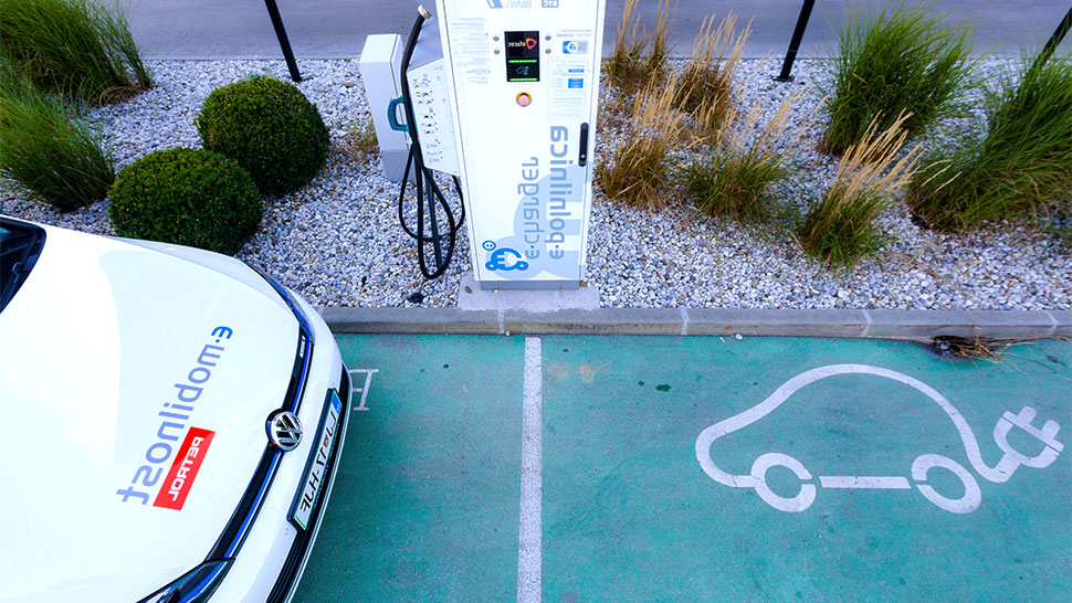 Na sliki je Petrolova e-polnilnica postavljena ob parkirnem prostoru, označenem z belo risbo električnega avtomobila in priključka. Na levi strani je parkiran avtomobil z oznako e-mobilnost Petrol, pogled pa je usmerjen od zgoraj navzdol proti polnilnemu mestu in okolici z okrasnim rastlinjem.