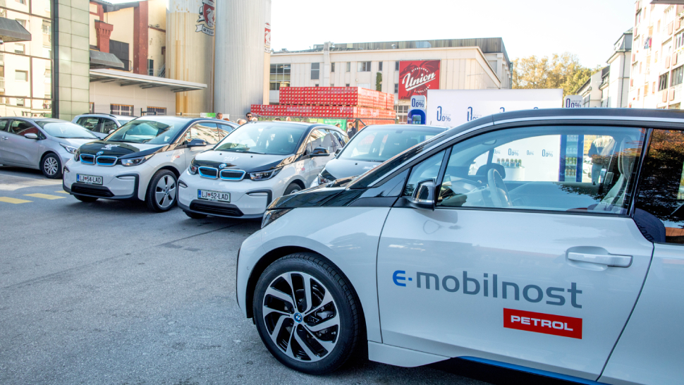 Na sliki je več električnih avtomobilov BMW i3 z oznako e-mobilnost Petrol, parkiranih na zunanjih parkirnih mestih v urbanem okolju. V ozadju je industrijska stavba z napisom »Union«.