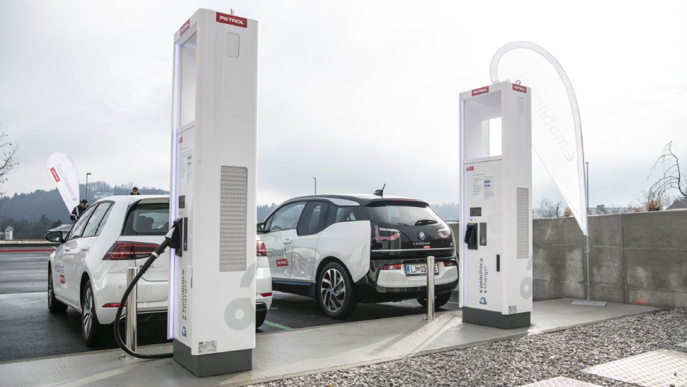 Na sliki sta dve električni vozili, Volkswagen Golf in BMW i3, ki se polnita na dveh Petrolovih polnilnih postajah ABB. Vozili sta parkirani na urejenem parkirišču, v ozadju pa je vidna zastava z napisom eMobilnost.