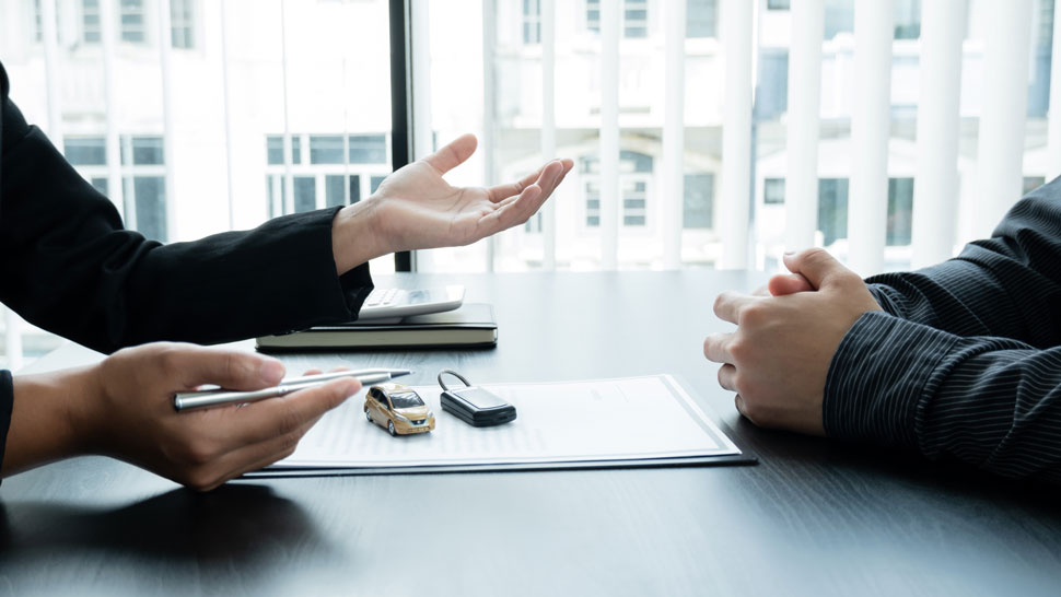 Na sliki sedita dve osebi za mizo, na kateri sta dokument in maketa avtomobila z avtomobilskim ključem. Ena oseba drži pisalo, druga pa ima roke prekrižane.