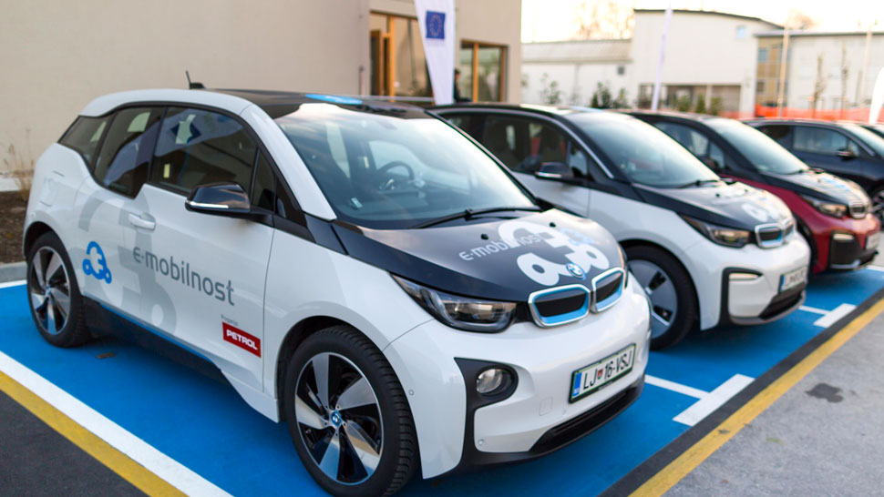 Na sliki je več električnih vozil, parkiranih na modro označenih parkirnih mestih. Vozila imajo napis »e-mobilnost Petrol« in so parkirana v vrsti pred stavbo.
