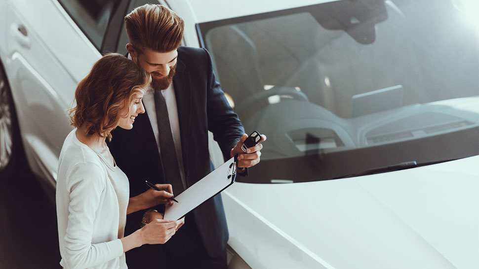 Na sliki sta moški in ženska, ki stojita ob belem avtomobilu. Ženska drži pisalo in podpisuje dokument na mapi, ki jo drži moški v poslovni obleki. Oba se nasmihata in zdita zadovoljna.