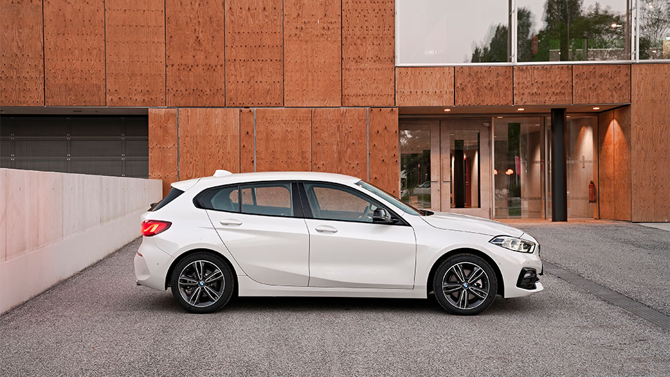 Na sliki je bel avtomobil, parkiran pred sodobno stavbo z lesenimi fasadnimi ploščami. Posnetek je narejen s strani, okolje pa deluje urbano in urejeno.